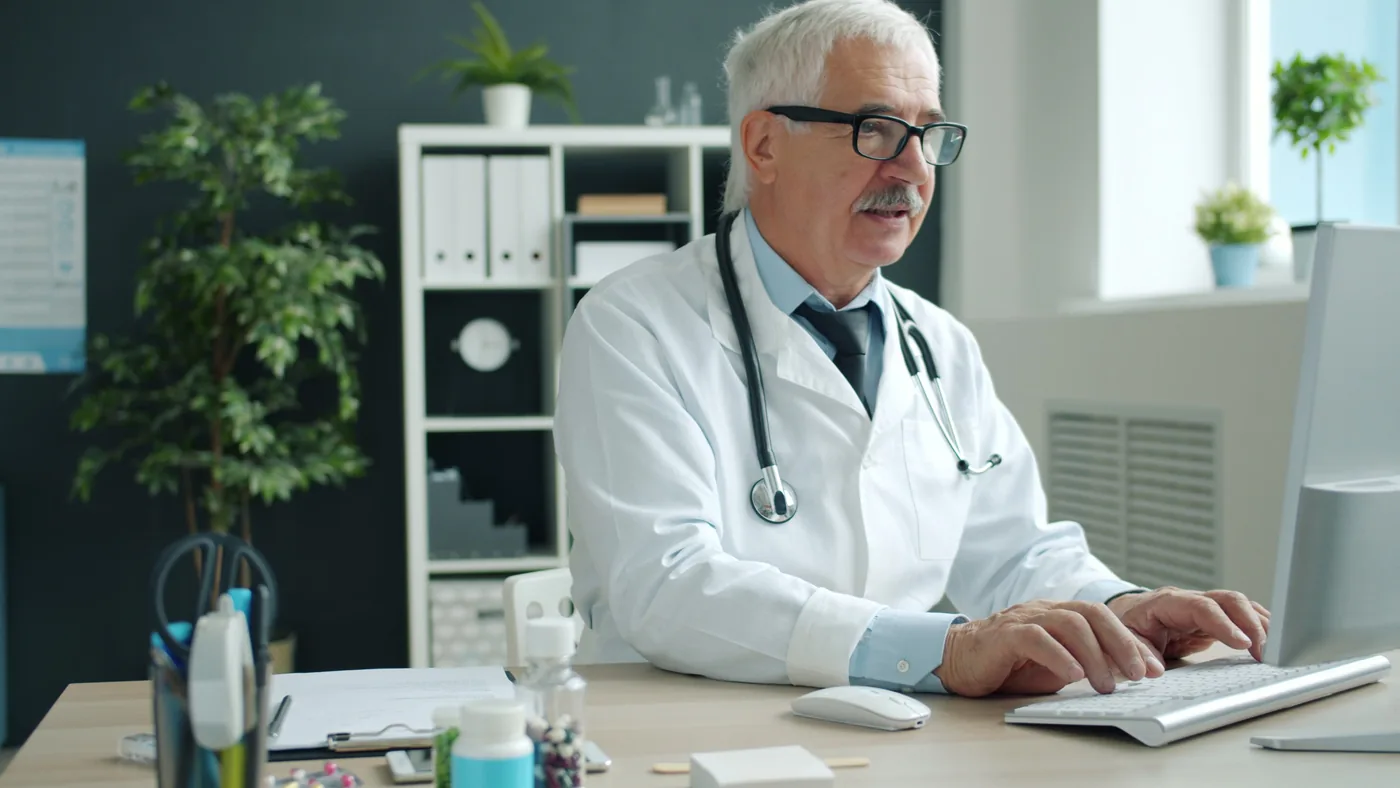 Ein Arzt mit weißem Haar, Brille und Stethoskop arbeitet am Computer in einer hellen Praxis.
